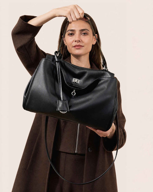 Woman in a brown coat holding the black leather flap bag by its long strap